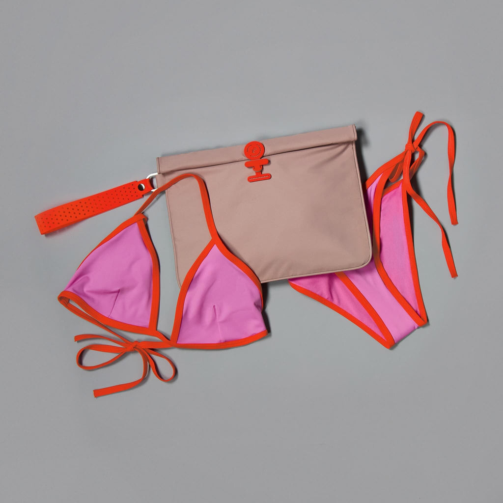 A coordinated swimwear and bag set laid flat on a gray background: a pink bikini top and matching bottoms with bright orange trim and ties, plus a beige pouch with orange trim and a flap closure.