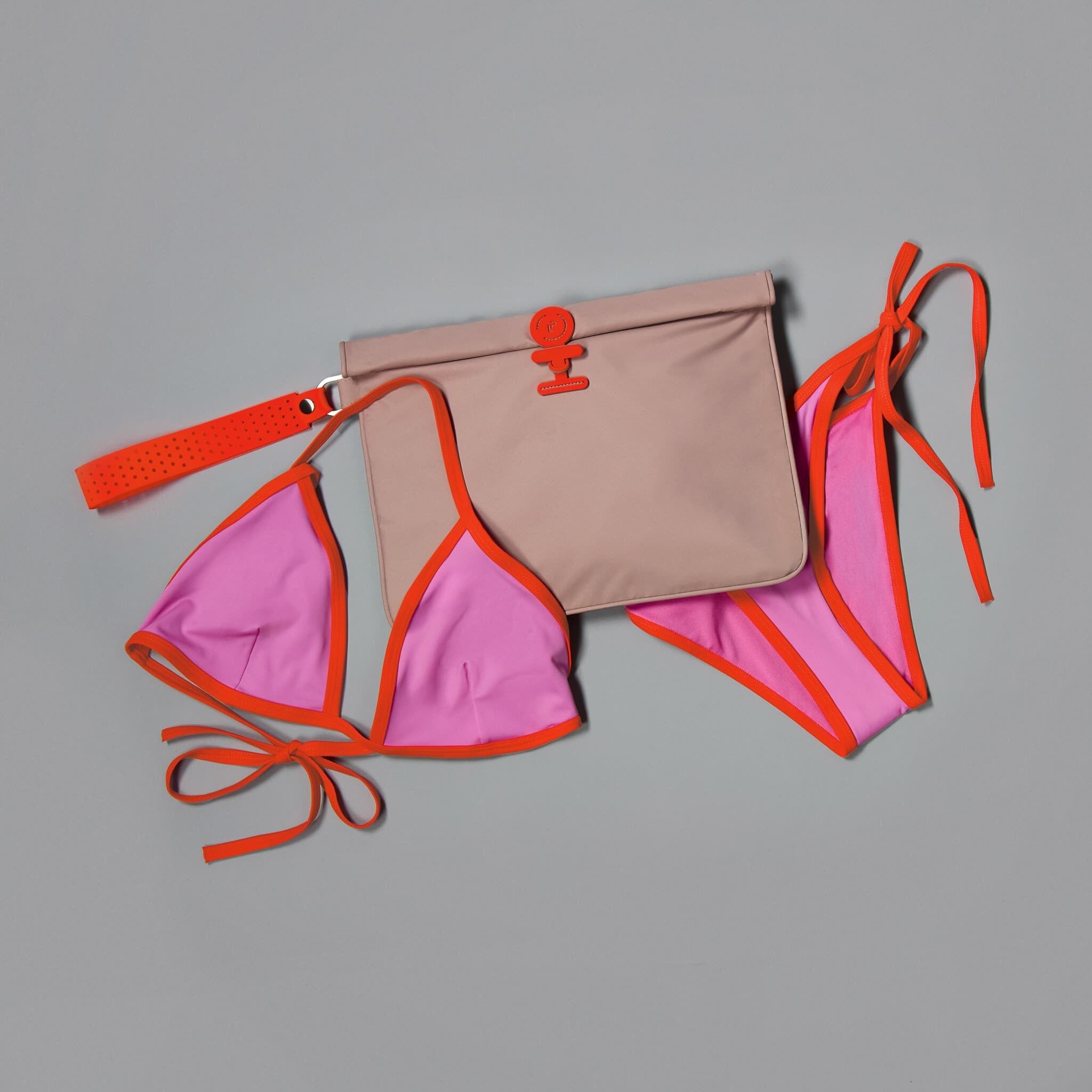 A coordinated swimwear and bag set laid flat on a gray background: a pink bikini top and matching bottoms with bright orange trim and ties, plus a beige pouch with orange trim and a flap closure.
